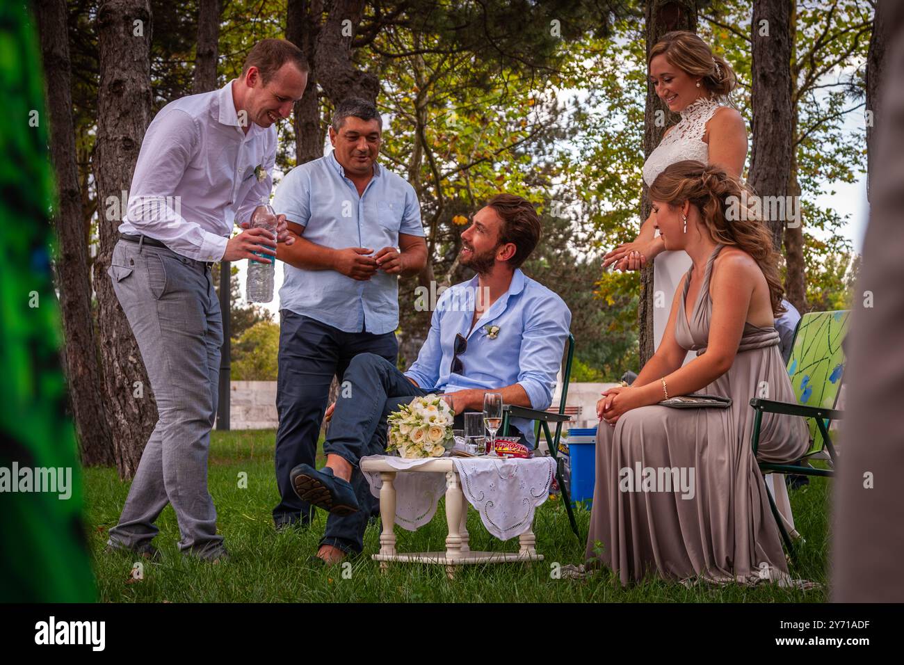 Due uomini portano bevande e dolci al testimone seduto a un piccolo tavolo festosamente apparecchiato, mentre la sposa è in fondo e la damigella d'onore guarda ridendo. Secondo la tradizione nuziale bulgara, la coppia serve il testimone e la damigella d'onore per tutta la giornata. Scattato a un matrimonio bulgaro a Dobrich, in Bulgaria. Foto Stock
