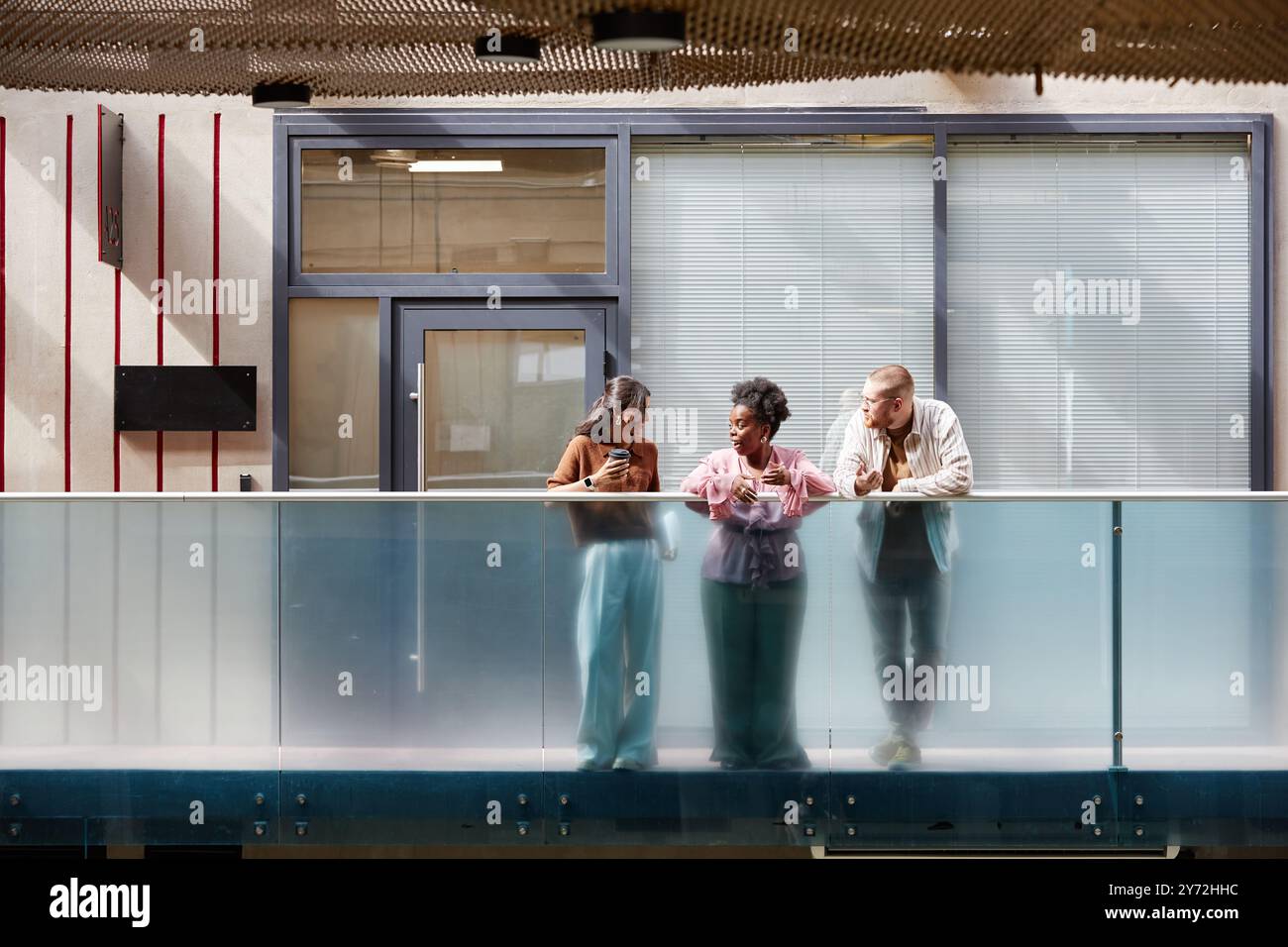 Riprese grandangolari di un gruppo di tre colleghi che chiacchierano allegramente durante la pausa mentre si alzavano sul balcone alla luce del sole che si proiettava attraverso il soffitto di vetro nel moderno business center, spazio per fotocopie Foto Stock