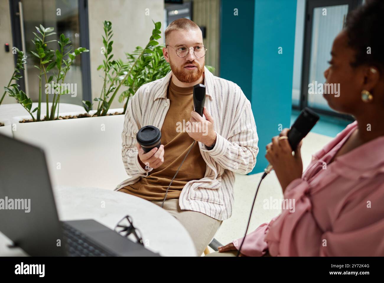 Ripresa media di un ospite podcast maschile che parla con la conduttrice mentre registra un nuovo episodio tenendo un microfono professionale seduto al tavolo del caffè, spazio per le copie Foto Stock