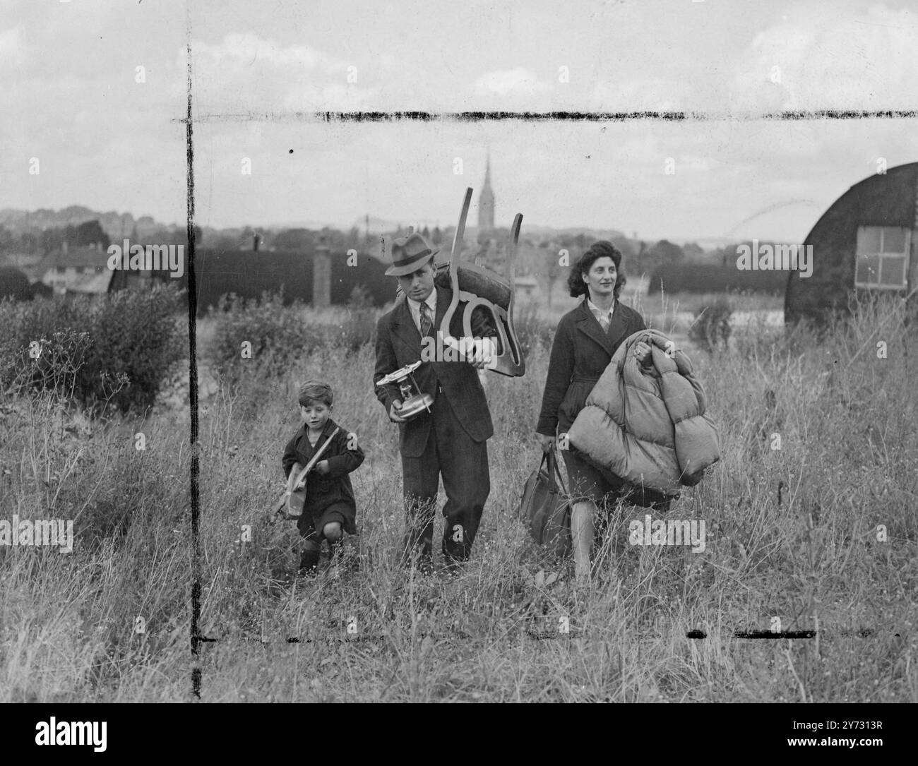 Gli squatters si muovono. Le famiglie senzatetto a Salsbury, Wilts, hanno risolto il problema da sole trasferendosi in una serie di capanne anti-esercito nella vicina West Harnham. L'Ufficio della Guerra e il Ministero della salute hanno deciso di non intraprendere alcuna azione contro di loro. Le immagini mostrano, il signor Norman Rose, un ex soldato, con 6 1/2 anni di servizio, visto con sua moglie e suo figlio Michael, che porta alcuni dei loro effetti personali dai "scavi" nelle vendite alla loro nuova casa nel sergente pasticcio a West Harnham. Sullo sfondo sorge la guglia della Cattedrale di Salisbury. 11 agosto 1946 Foto Stock