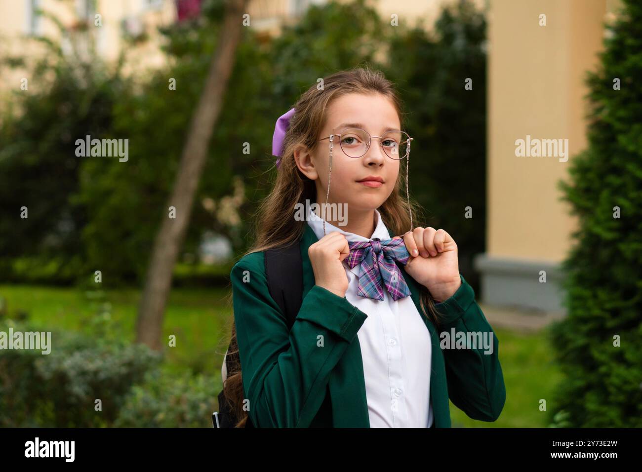 Una giovane ragazza con un blazer verde che regola il suo papillon mentre è in piedi in un parco, guardando direttamente la telecamera con un'espressione seria. Foto Stock