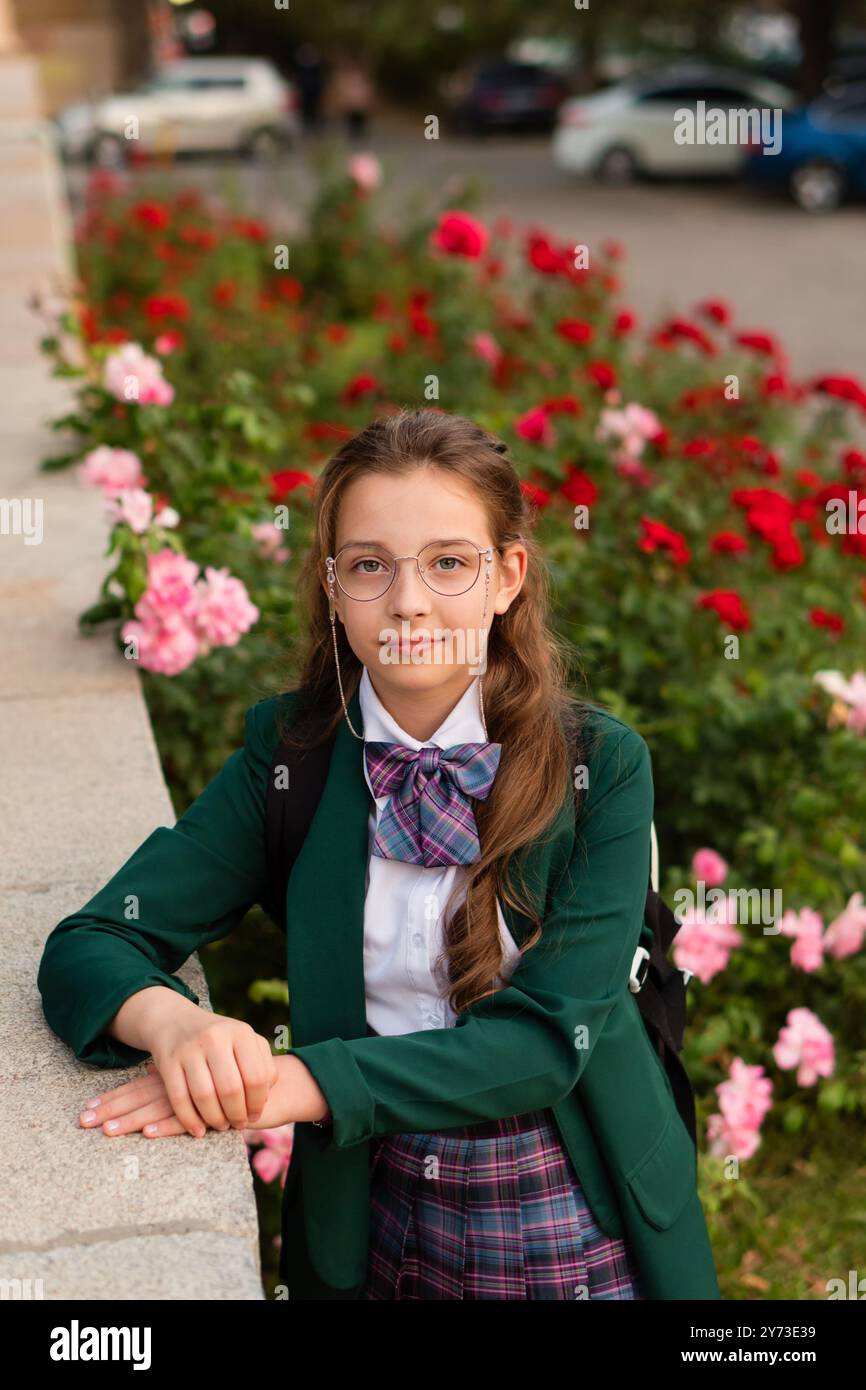 Ragazza in uniforme scolastica in piedi vicino ai fiori, che posa ponderatamente con un sorriso morbido. Ritratto di strada di una studentessa Foto Stock