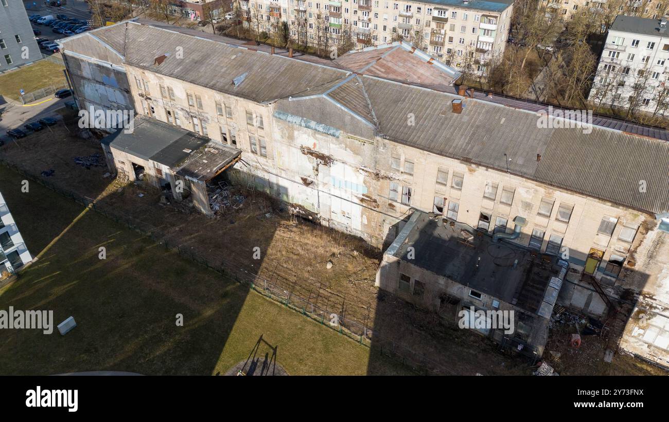 Una vista aerea di un edificio fatiscente circondato da erba e detriti sovrastati. Nelle vicinanze si trovano edifici residenziali sullo sfondo, a contrasto Foto Stock