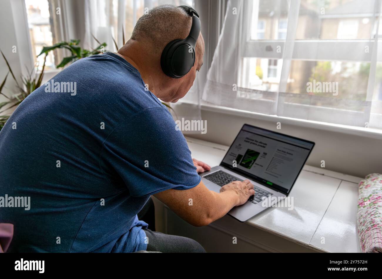 Un uomo anziano a casa con cuffie wireless e un computer portatile, che impara e cerca di tenersi aggiornato con la tecnologia moderna. Una tecnologia usa con Foto Stock