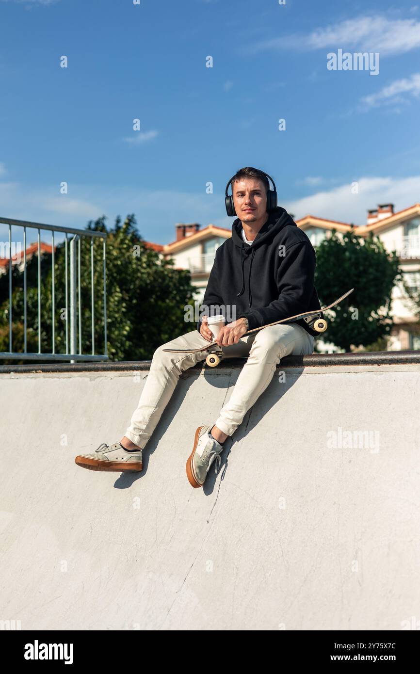 pattinatore verticale contemplativo seduto in uno skate park, sorseggiando una tazza di caffè caldo mentre ascolti la musica attraverso le cuffie. Con uno sguardo lontano, Foto Stock