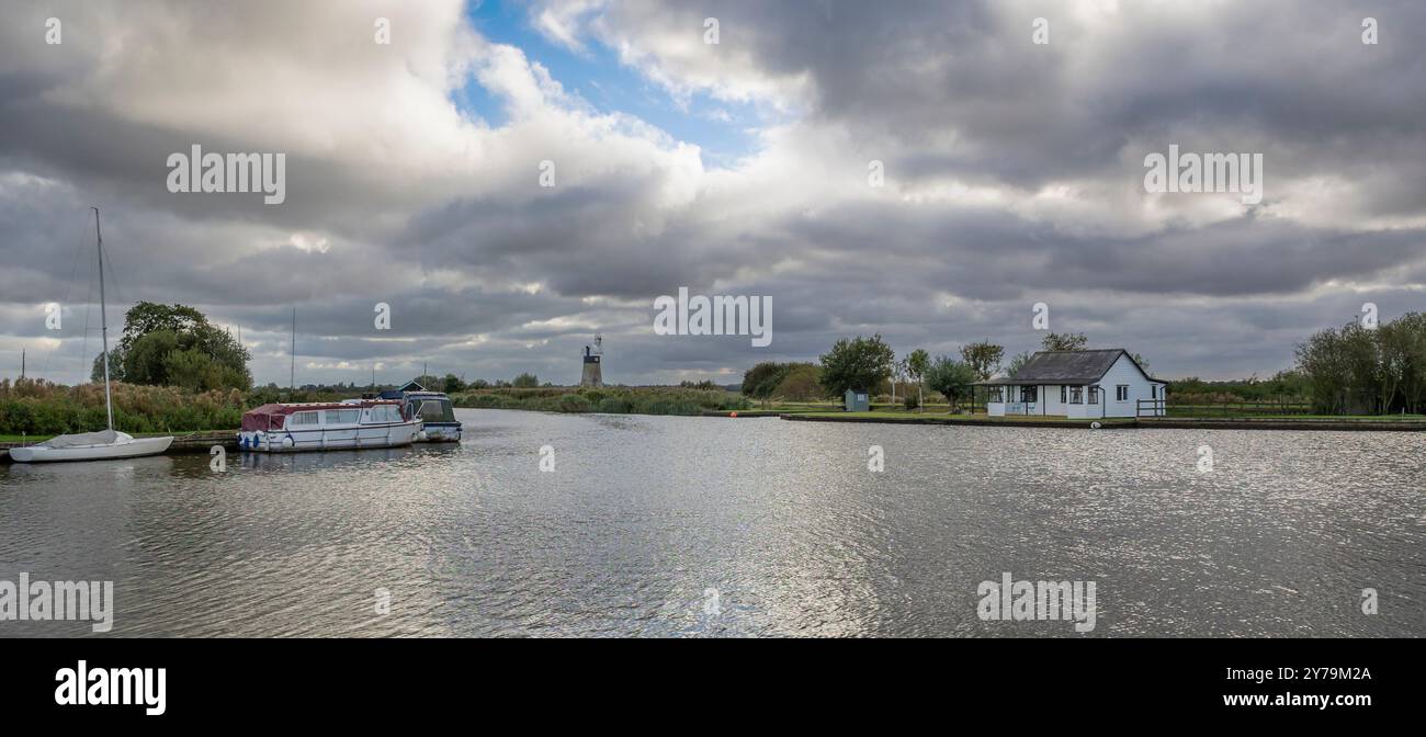 Una veduta dei norfolk broad presa a Thurne. La foto mostra una piccola casa vicino al fiume e un mulino a vento in lontananza. piccole barche sono ormeggiate Foto Stock