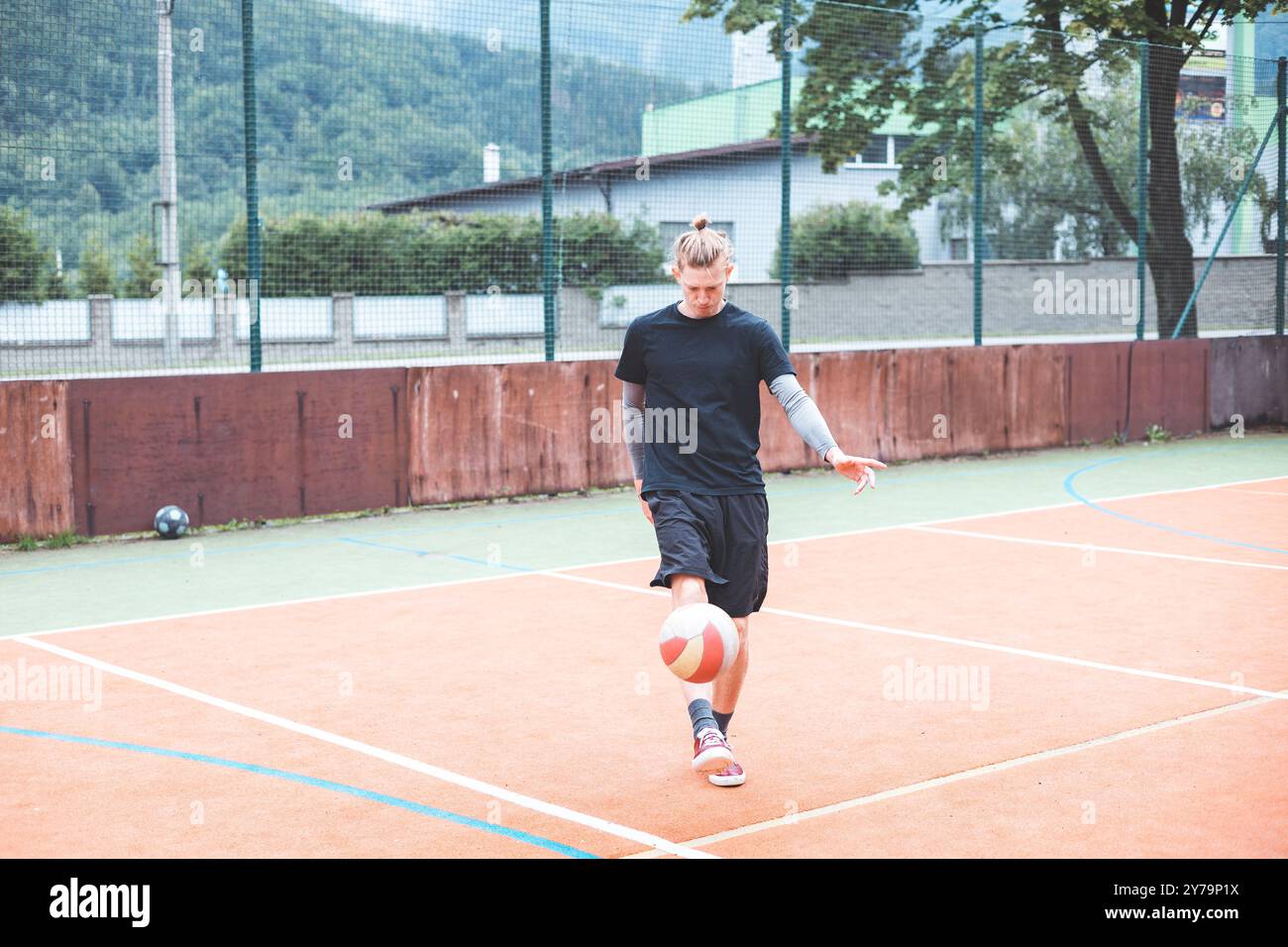 Il giovane con un uomo Bun giocherà una partita di calcio su un vivace campo arancione durante una giornata di sole. Indossando un abbigliamento sportivo casual, si concentra sul controllo del Bal Foto Stock