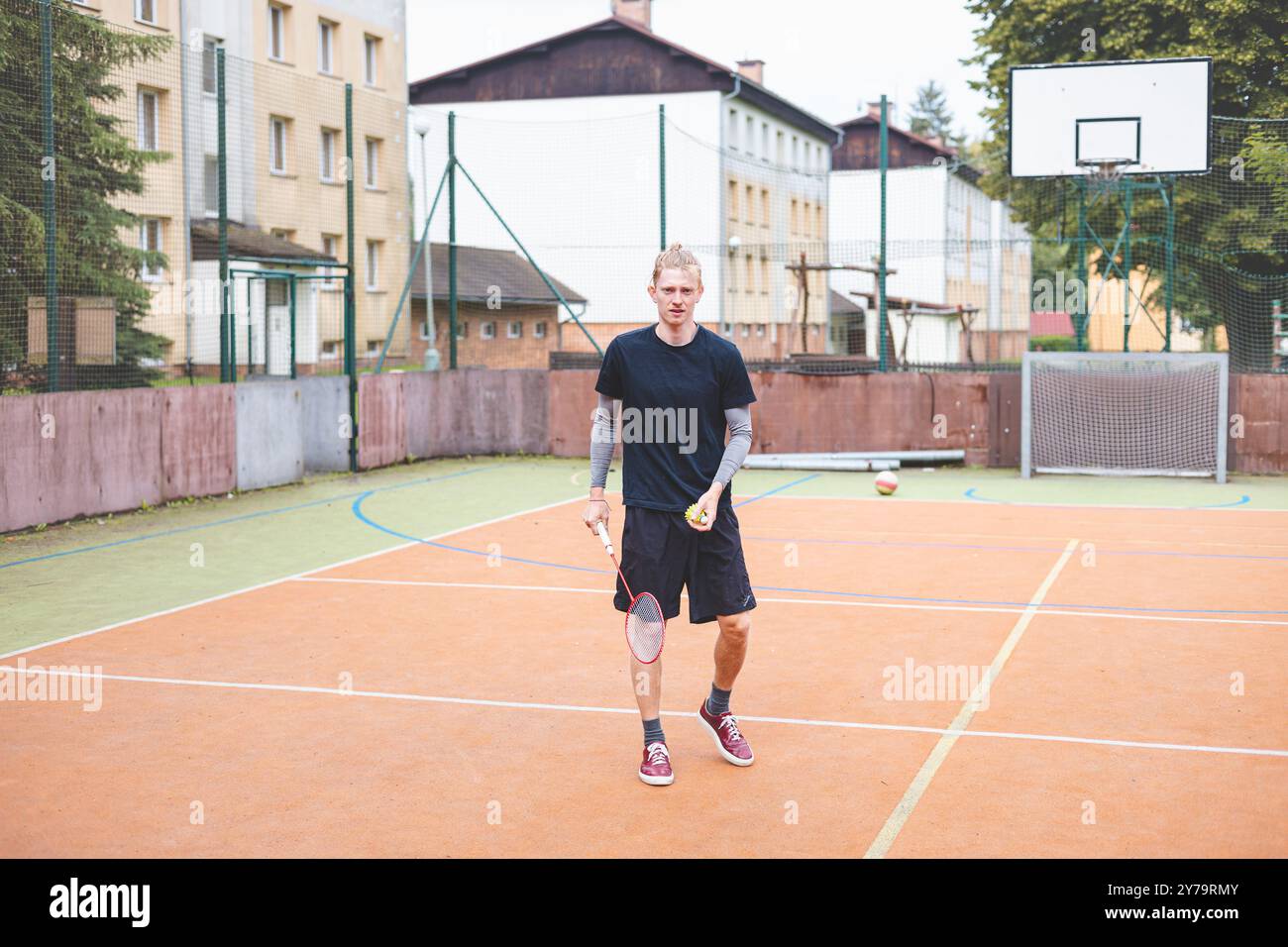 Il giocatore di badminton prende un rubinetto durante una partita su un campo all'aperto. Il momento informale si svolge in un ambiente urbano, con edifici circostanti Foto Stock Il giocatore di badminton prende un rubinetto durante una partita su un campo all'aperto. Il momento informale si svolge in un ambiente urbano, con edifici circostanti Foto Stock