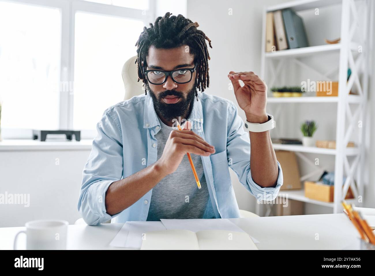 Giovane uomo africano intelligente in occhiali che dice qualcosa e guarda la macchina fotografica mentre lavora in casa Foto Stock
