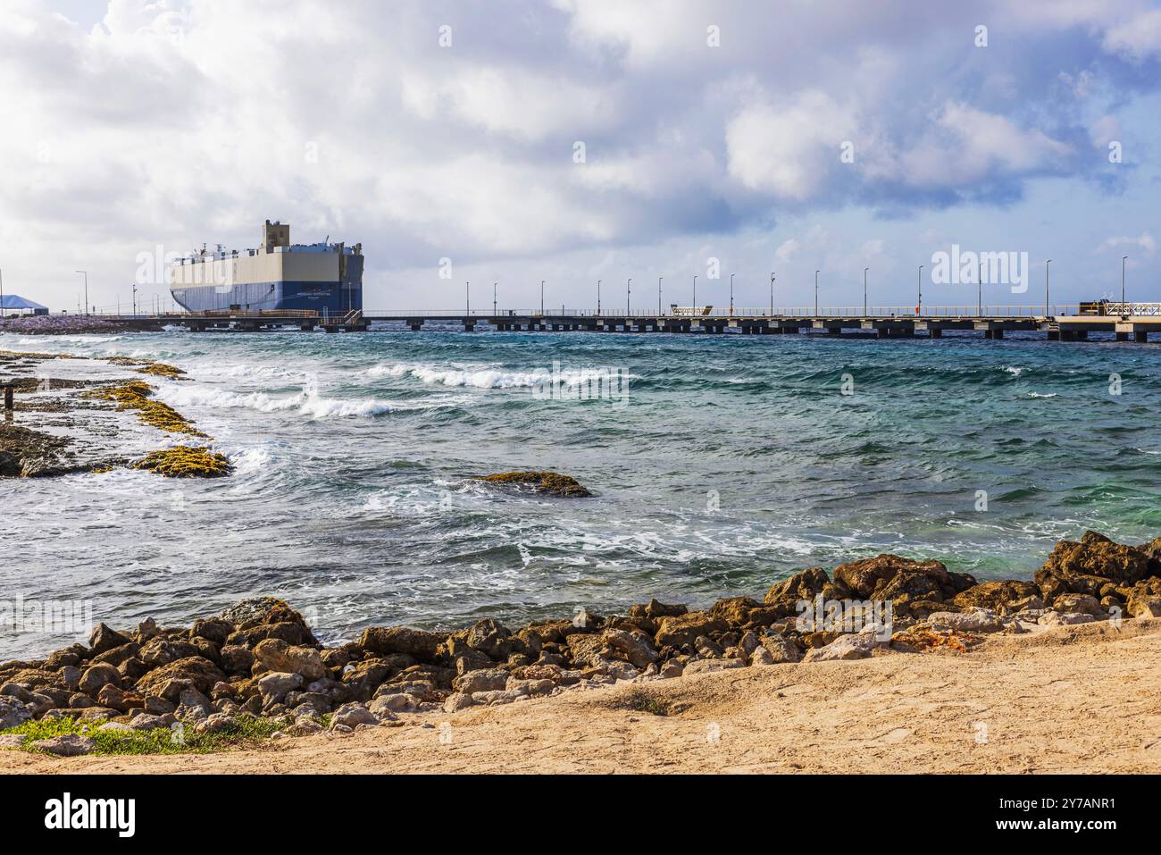 Nave da carico attraccata su un molo con onde che colpiscono la costa rocciosa e cieli nuvolosi, situata sulla costa dell'isola di Curacao nel Mar dei Caraibi. Foto Stock