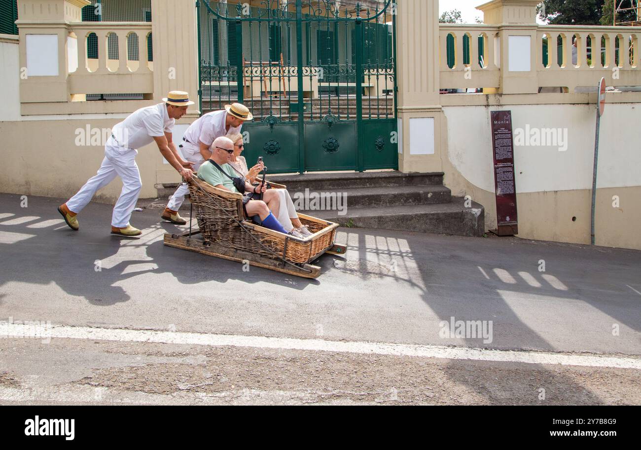 Turisti e turisti che si divertono facendo un giro sulle tradizionali slitte da slittino con cesto di vimini a Monte, Funchal, sull'isola portoghese di Madeira Foto Stock