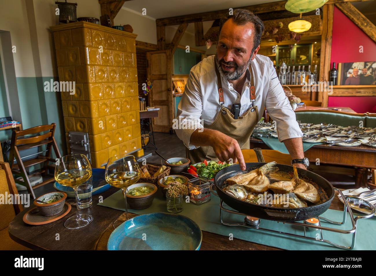 René Bobzin posa la padella di pesce fritto su un supporto termico al tavolo festosamente apparecchiato, circondato da vino bianco, insalata e piccole ciotole di antipasti. Morgenitz, comune di Mellenthin, Meclemburgo-Pomerania Anteriore, Germania. Foto Stock