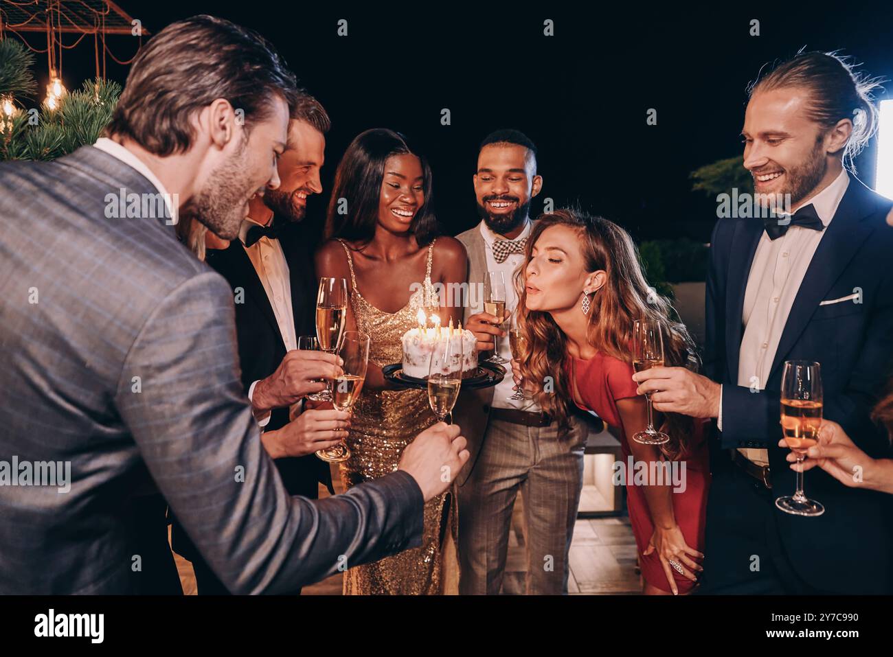 Bella giovane donna con un abito da sera che soffia candele sulla torta mentre festeggia il compleanno con gli amici Foto Stock