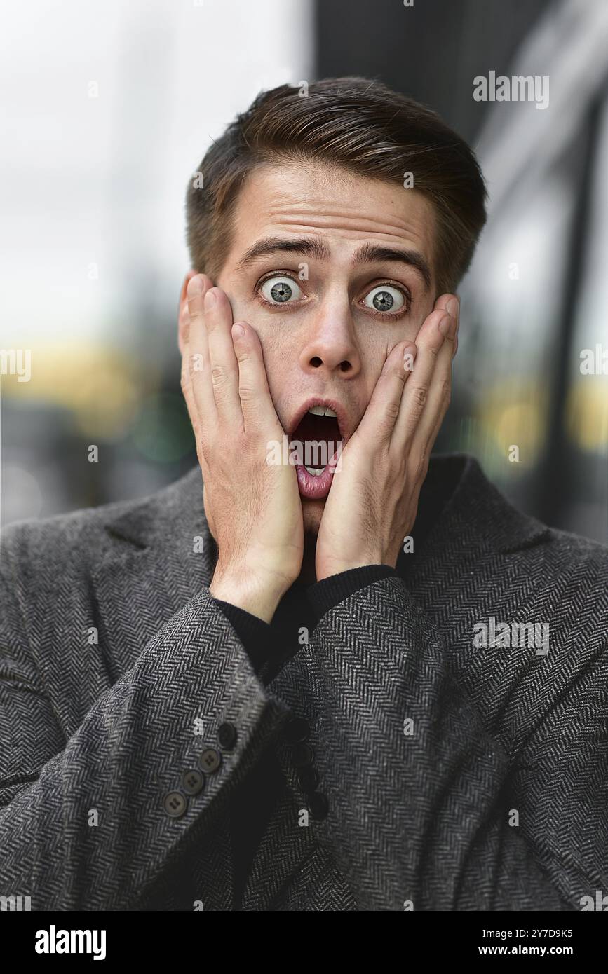 Sorpreso giovane uomo caucasico di successo in piedi all'aperto. Uomo che indossa un cappotto grigio e un maglione blu Foto Stock