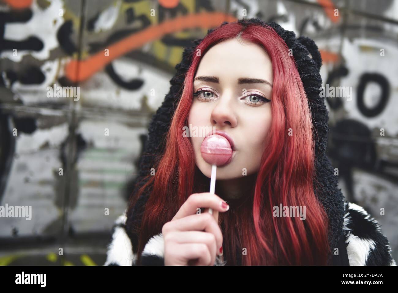 Ritratto di una giovane bellezza in una felpa con cappuccio con un delizioso lollipop rosa, in posa contro un muro di graffiti. Emozioni positive. Stile Urrban Foto Stock