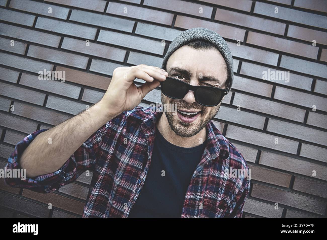 Un giovane adulto che indossa occhiali da sole in piedi su un muro di mattoni marroni sorridente con la faccia felice che strizza l'occhio alla telecamera Foto Stock