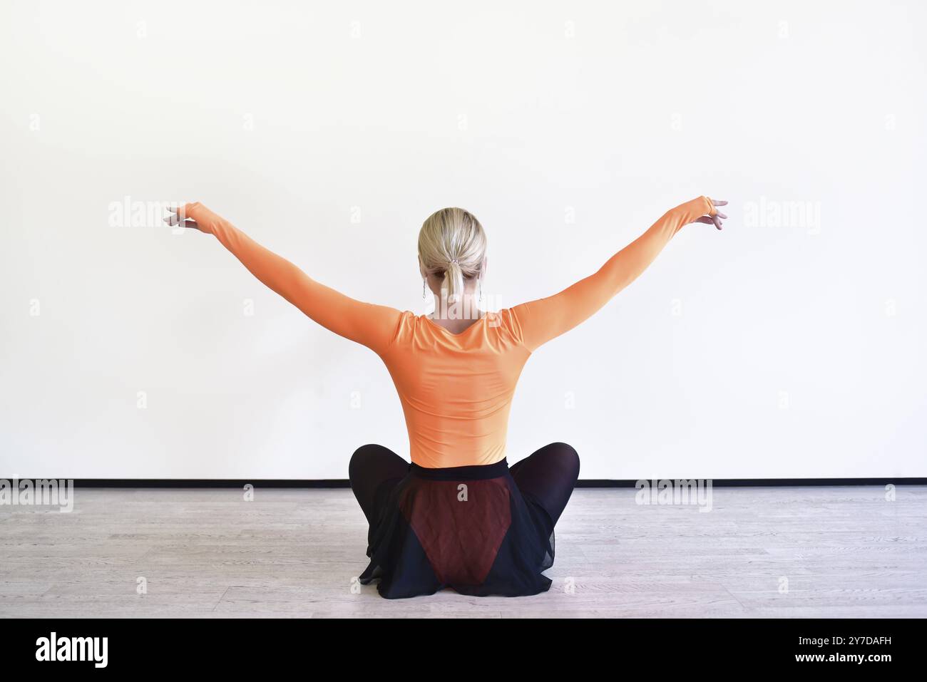La vista posteriore di un'affascinante ballerina bionda è seduta sul pavimento nella sala da ballo per riscaldarsi prima dello spettacolo Foto Stock