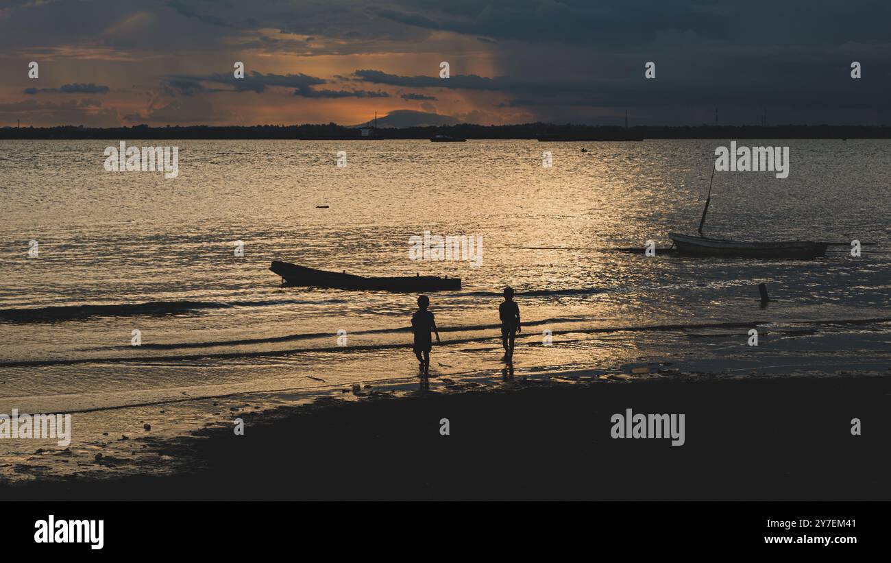Una foto drammatica di due ragazzi che camminano su una spiaggia al tramonto, con le loro silhouette incorniciate contro un cielo ardente. Foto Stock