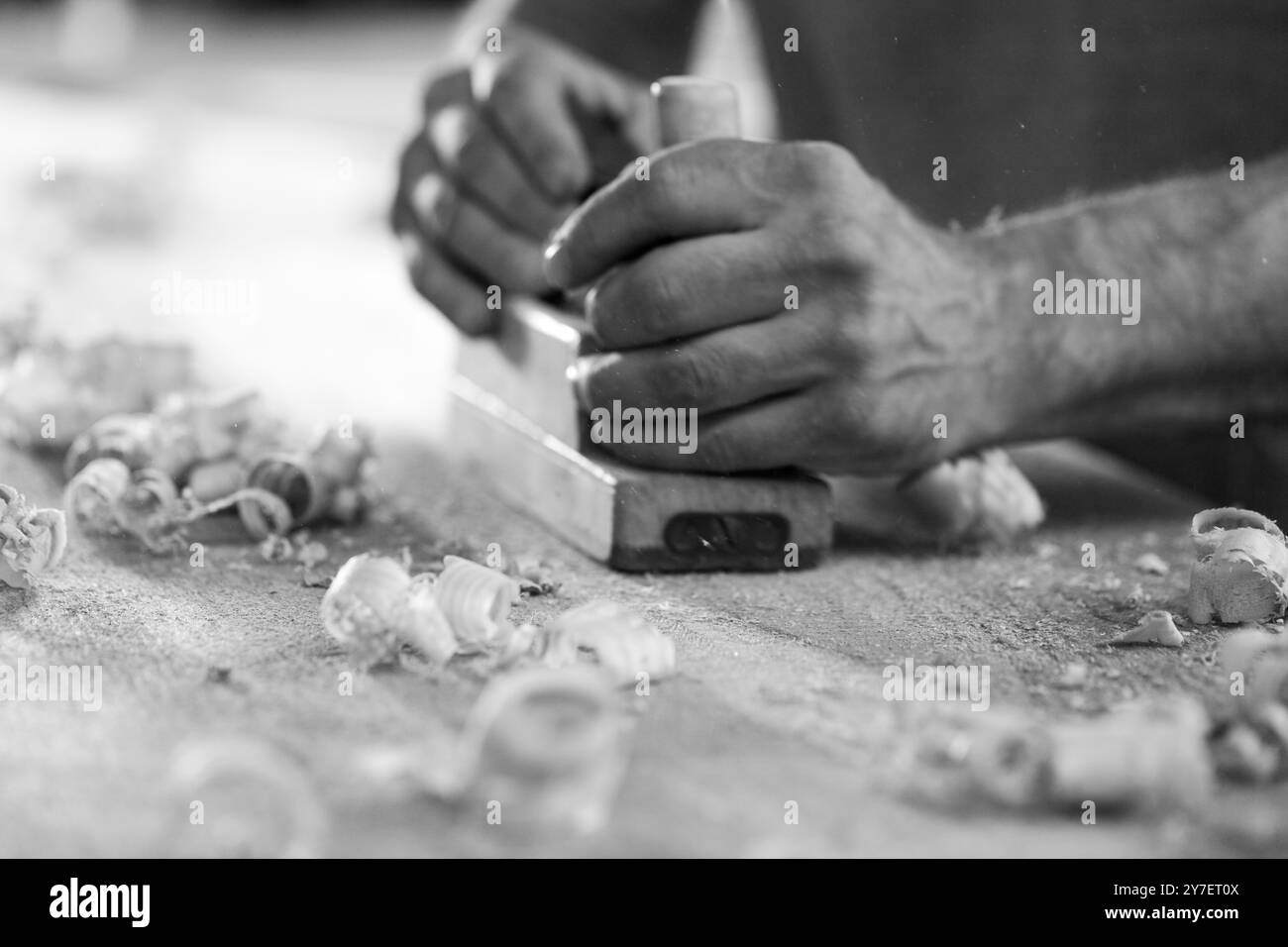 Carpenter sta levigando una tavola di legno con una pialla nella sua officina, circondata da trucioli di legno. - nero n bianco - Foto Stock
