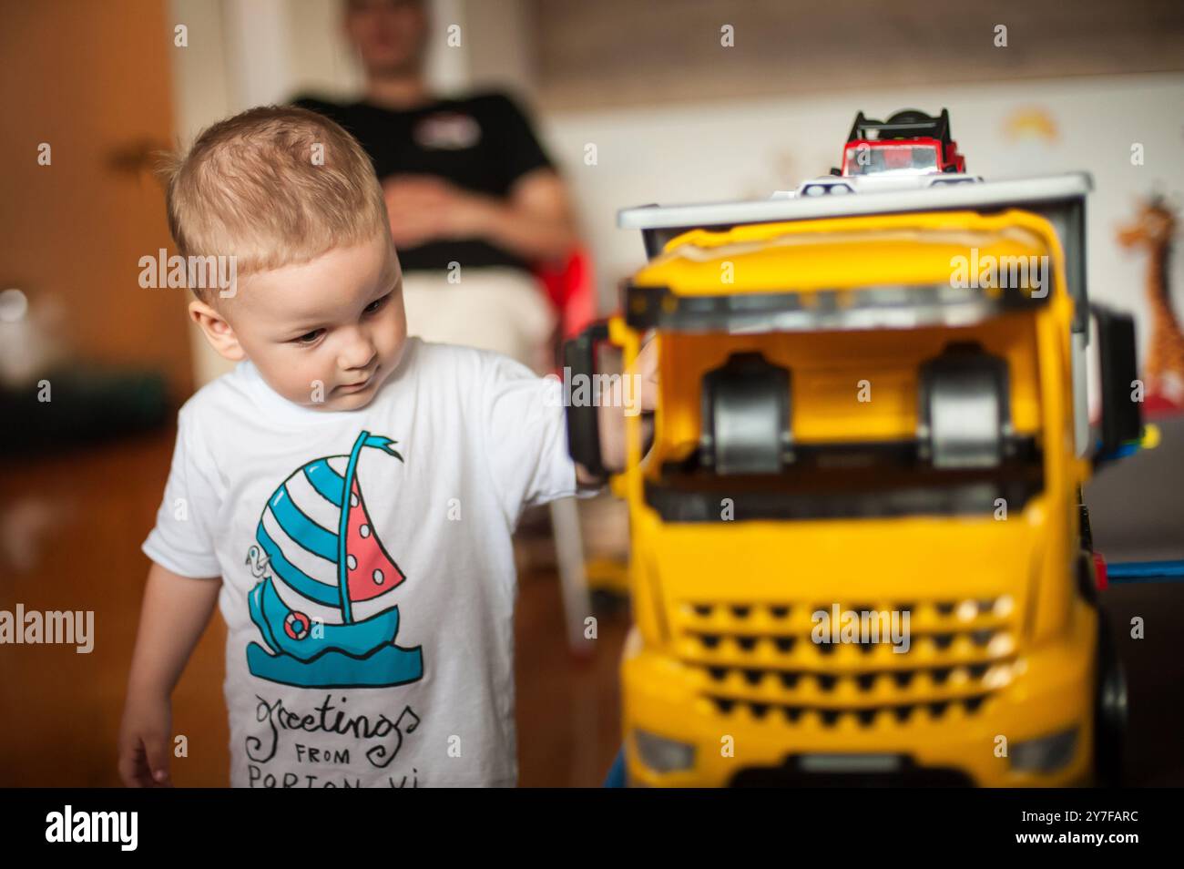 Un bambino esamina un grande camion giocattolo giallo con curiosità, circondato da giocattoli colorati in un soggiorno caldo e invitante, mentre un genitore si rilassa in t Foto Stock