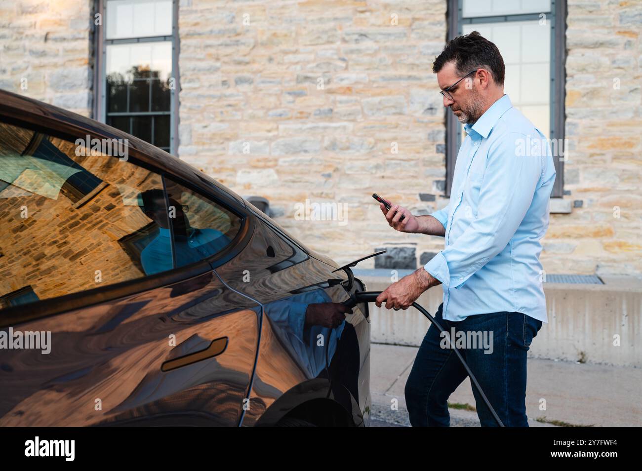 Uomo che utilizza lo smartphone durante la ricarica di un veicolo elettrico EV all'esterno. Foto Stock