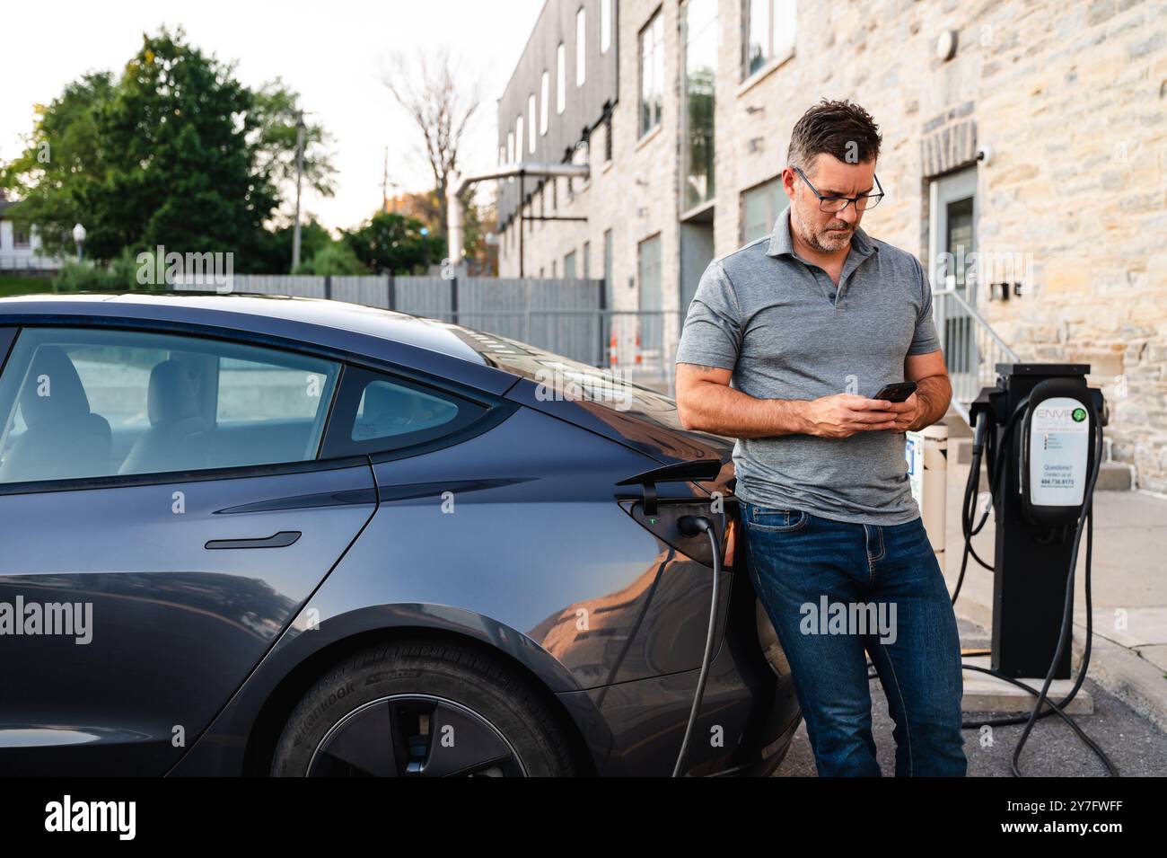 Uomo che utilizza il telefono mentre aspetta che l'auto EV si ricarichi presso una stazione esterna. Foto Stock