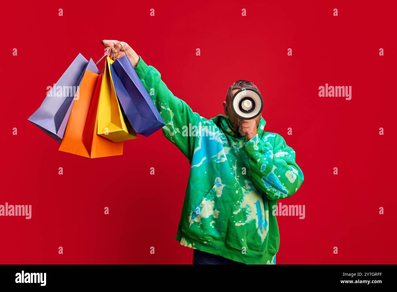 Uomo in elegante felpa con cappuccio che tiene in mano borse colorate e parla in megafono su sfondo rosso. Offerte a tempo limitato Foto Stock