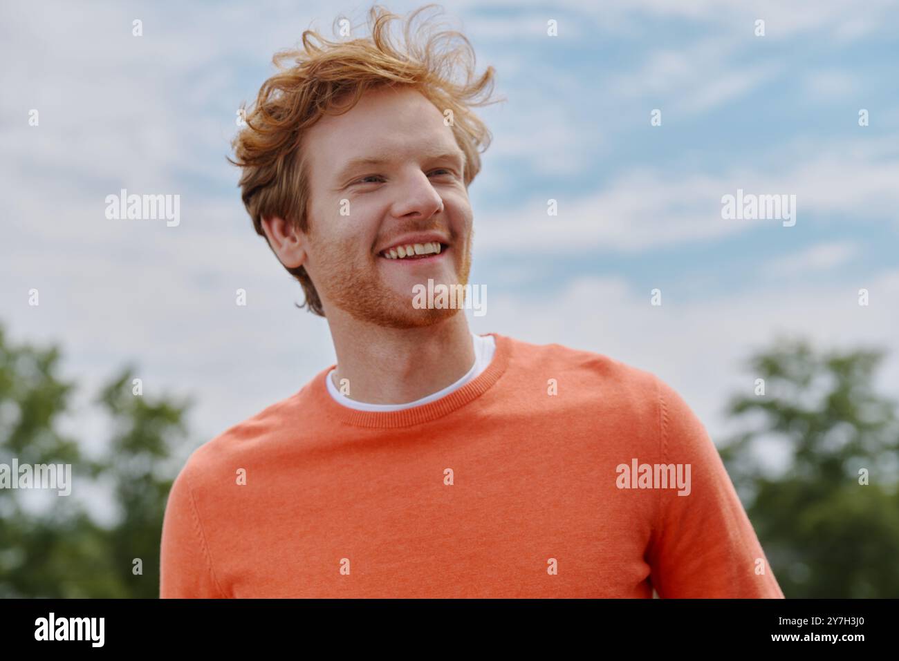 Allegro giovane uomo di capelli rossi che guarda lontano e sorride mentre è in piedi all'aperto Foto Stock