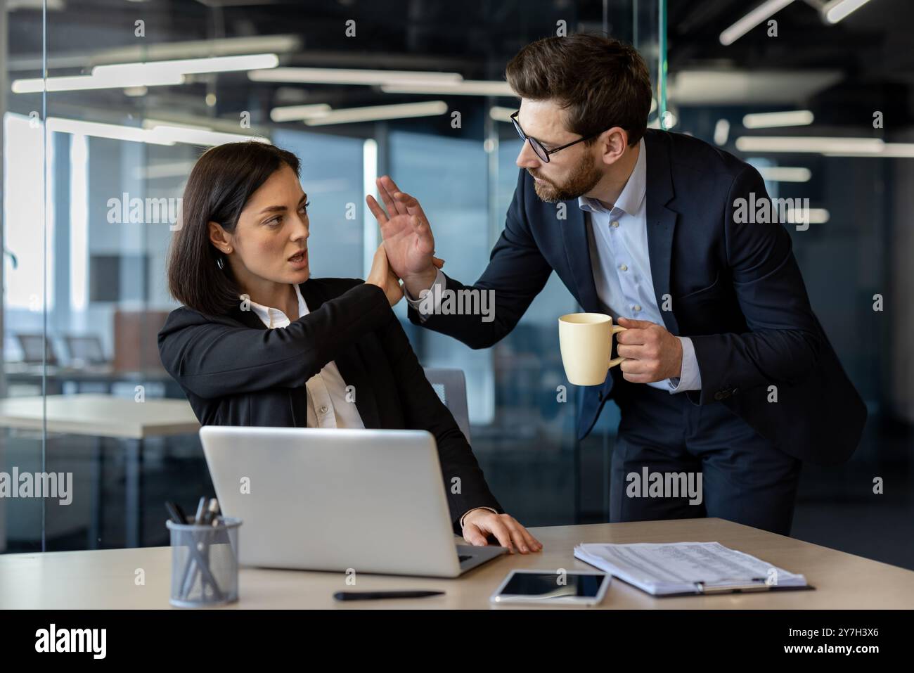 Una donna d'affari seduta alla scrivania utilizza un linguaggio del corpo forte per fermare l'avanzamento indesiderato del collega con una tazza di caffè, illustrando le dinamiche del luogo di lavoro, i conflitti e la prevenzione delle molestie. Foto Stock