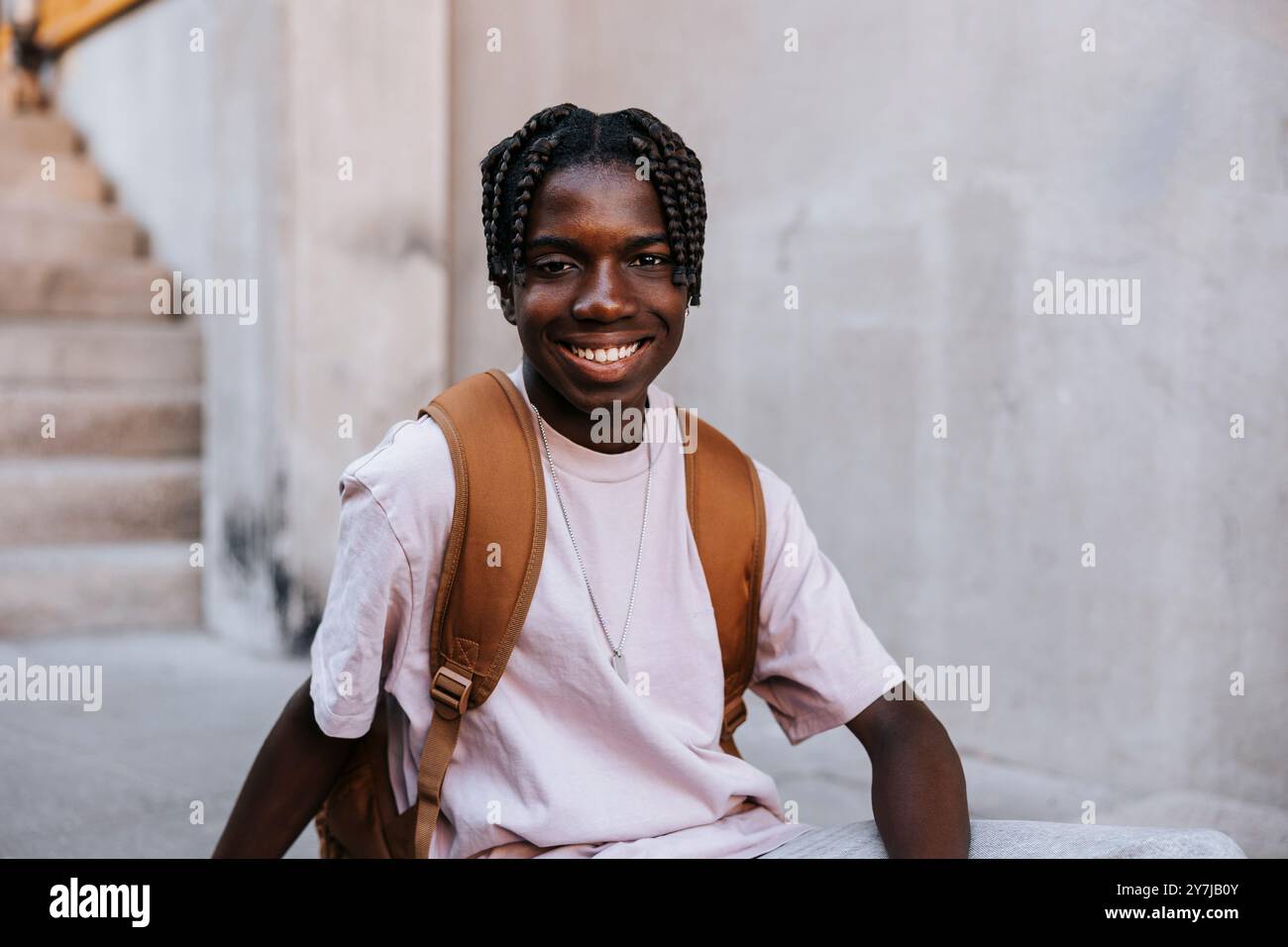 Ritratto di uno studente maschio sorridente con capelli intrecciati seduto vicino al muro dell'edificio scolastico Foto Stock