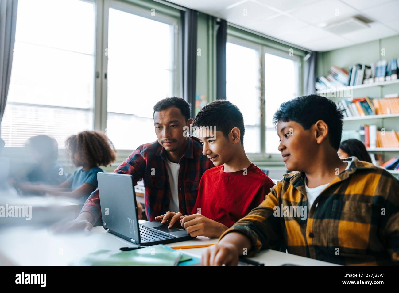 Insegnante di sesso maschile che assiste gli studenti che studiano con un computer portatile in classe alla scuola elementare Foto Stock