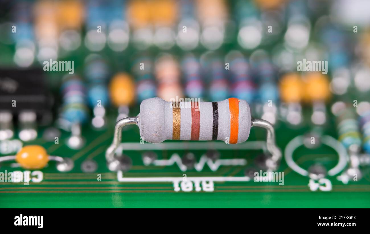 Primo piano di una resistenza bianca con codice colorato standard su una scheda a circuito stampato verde. Componente elettronico con sistema a banda colorata su circuito stampato sfocatura con bokeh. Foto Stock