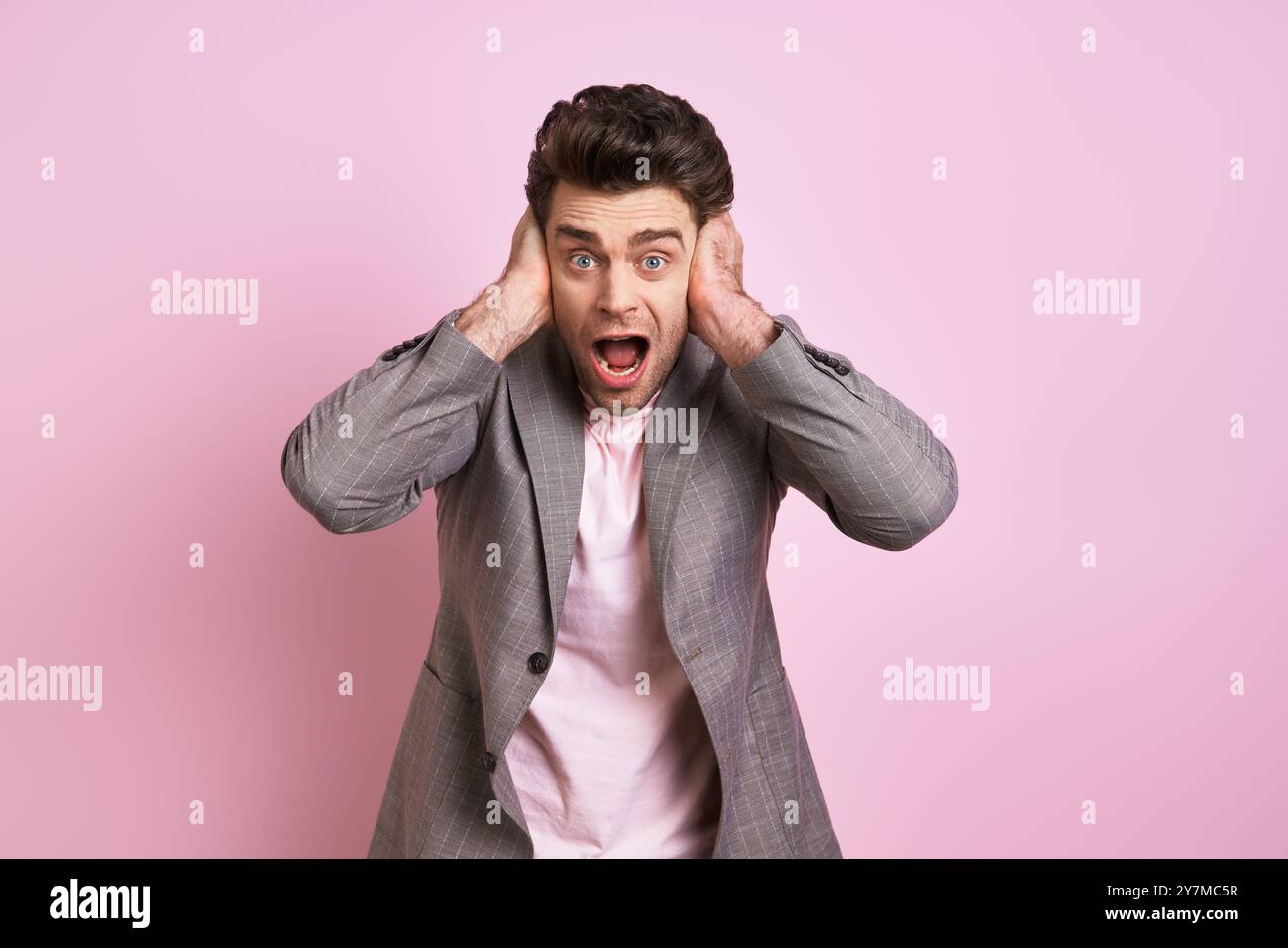 Un giovane scioccato che copre le orecchie con le mani mentre si trova sullo sfondo rosa Foto Stock Un giovane scioccato che copre le orecchie con le mani mentre si trova sullo sfondo rosa Foto Stock