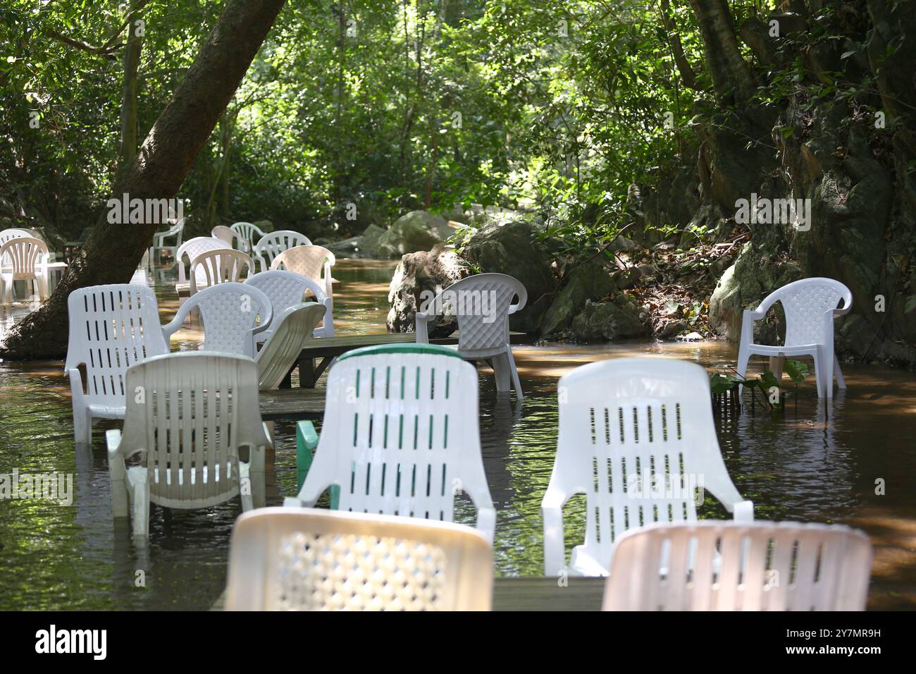 Sedie vuote di plastica bianca sono collocate nel torrente di un caffè ai piedi di una collina con una cascata che scorre attraverso di essa. Foto Stock