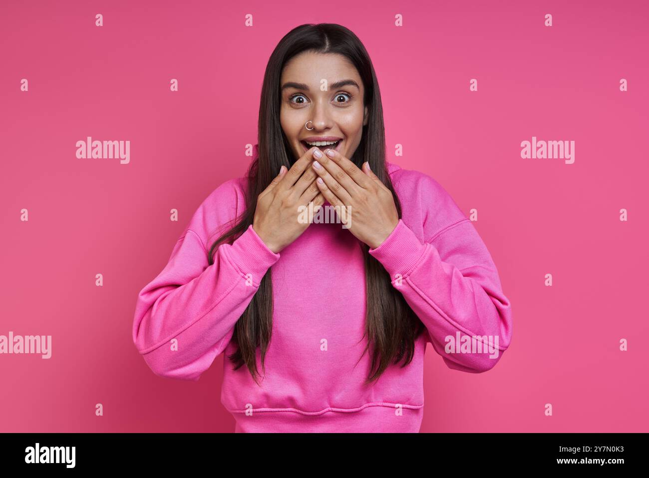 Una giovane donna sorpresa con una camicia con cappuccio che copre la bocca con le mani su sfondo rosa Foto Stock Una giovane donna sorpresa con una camicia con cappuccio che copre la bocca con le mani su sfondo rosa Foto Stock