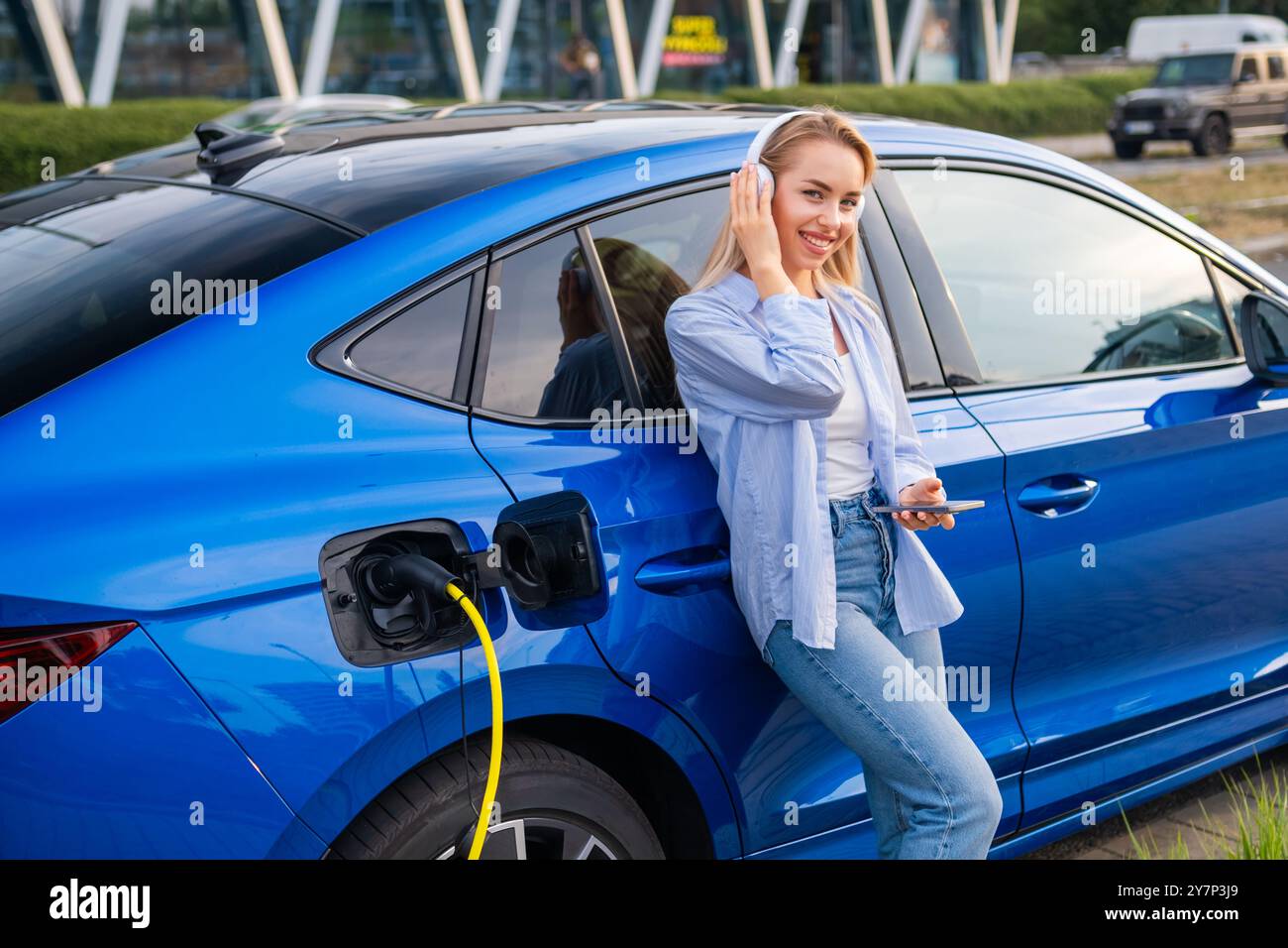 Donna monitora lo stato di carica delle sue auto elettriche utilizzando il suo smartphone, mentre indossa le cuffie in piedi vicino all'auto blu. Foto Stock