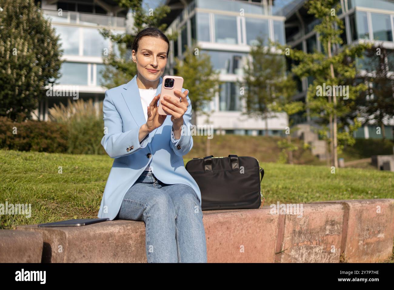 Una donna d'affari utilizza uno smartphone mentre lavora in remoto, approfittando del clima caldo e del sole per lavorare all'aperto in un parco urbano Foto Stock