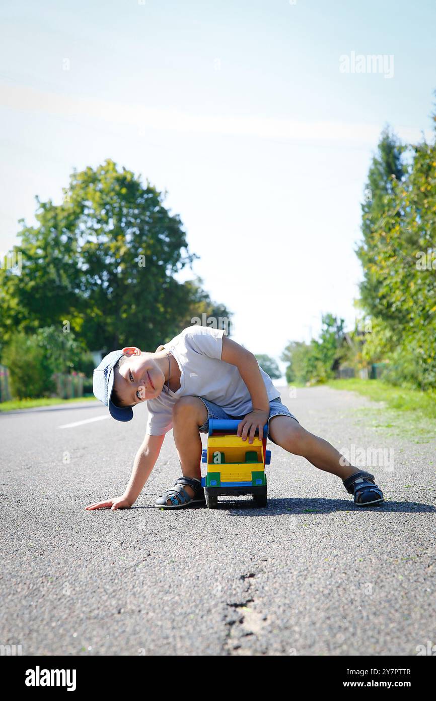 Un bambino in età prescolare gioca su una strada asfaltata con una grande macchinetta giocattolo in una soleggiata giornata estiva Foto Stock