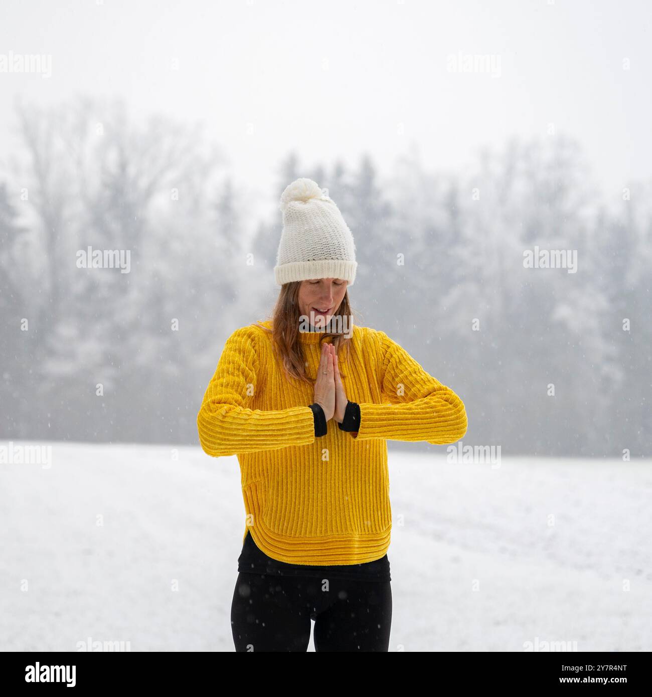 Giovane donna spirituale con un cappello e un maglione giallo brillante in piedi nel mezzo della tranquilla natura invernale fredda che prega o pratica gratitudine e m Foto Stock