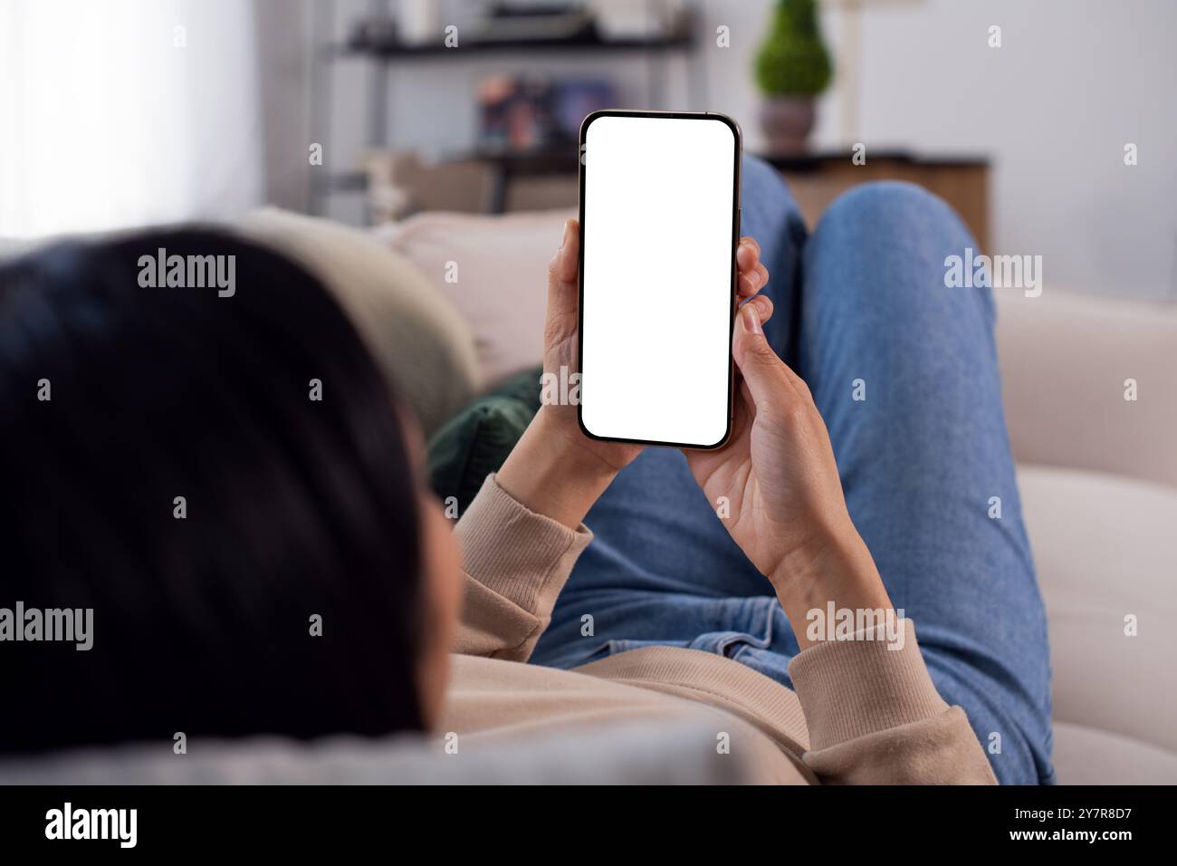 Una giovane donna asiatica irriconoscibile che regge un mockup di smartphone con uno schermo bianco vuoto che si trova sull'accogliente divano di casa, punto di vista Foto Stock