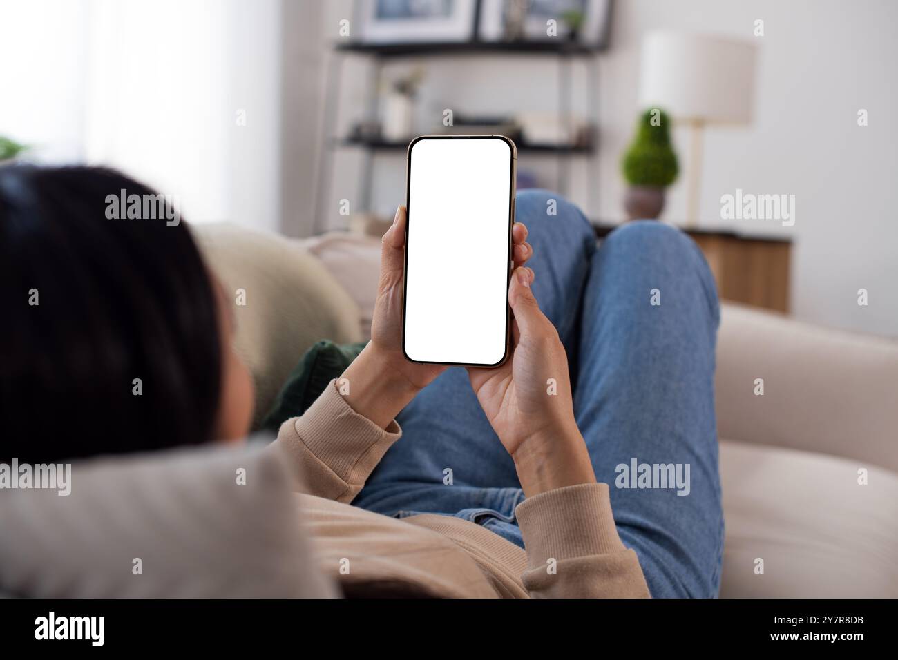 Una giovane donna asiatica irriconoscibile che regge un mockup di smartphone con uno schermo bianco vuoto che si trova sull'accogliente divano di casa, punto di vista Foto Stock