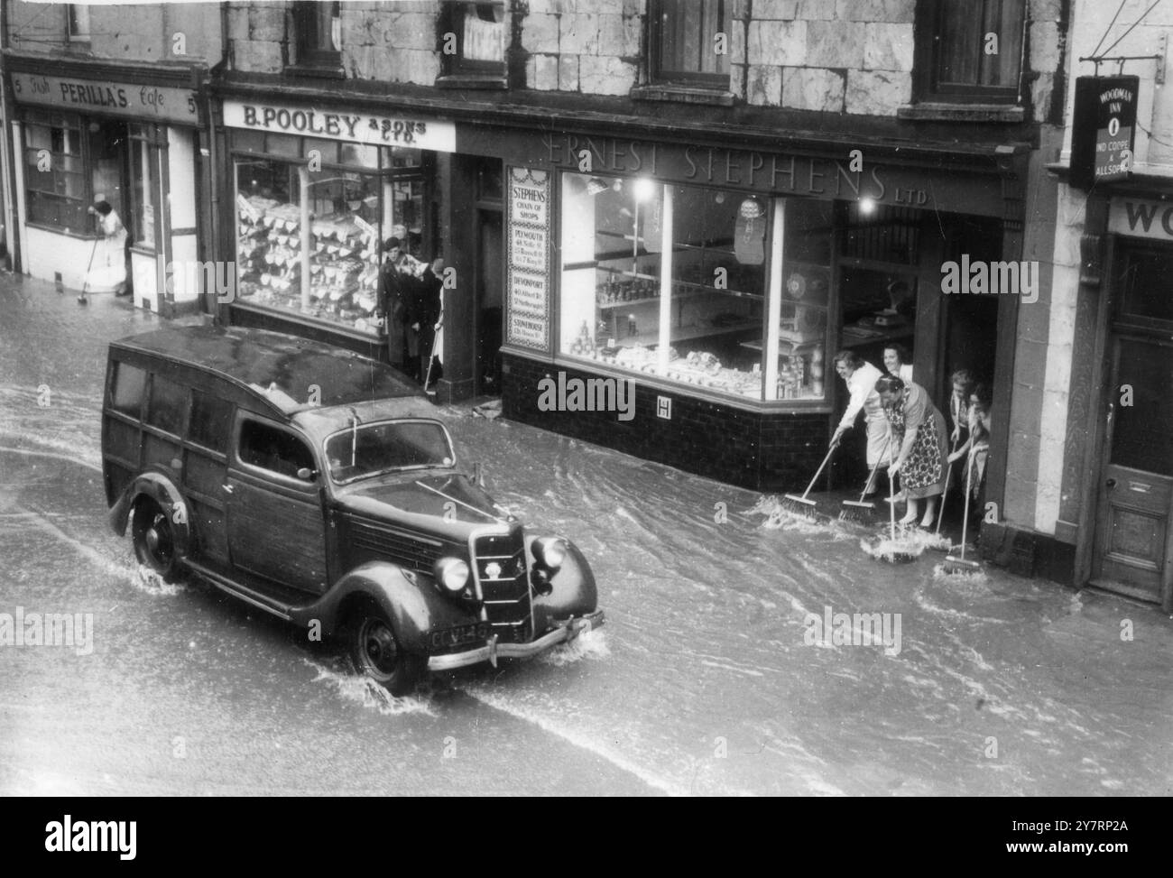 LE INONDAZIONI HANNO COLPITO PLYMOUTH. Plymouth, Devonshire: Le donne usano le scope per tenere l'acqua fuori dai negozi di Plymouth's Main Street, mentre un'auto passa durante le inondazioni che hanno seguito le piogge torrenziali. Una volta la pioggia stava cadendo a un ritmo di circa due centimetri all'ora. I vigili del fuoco hanno ricevuto 55 chiamate di emergenza da edifici allagati in mezz'ora 16 agosto 1952 Foto Stock