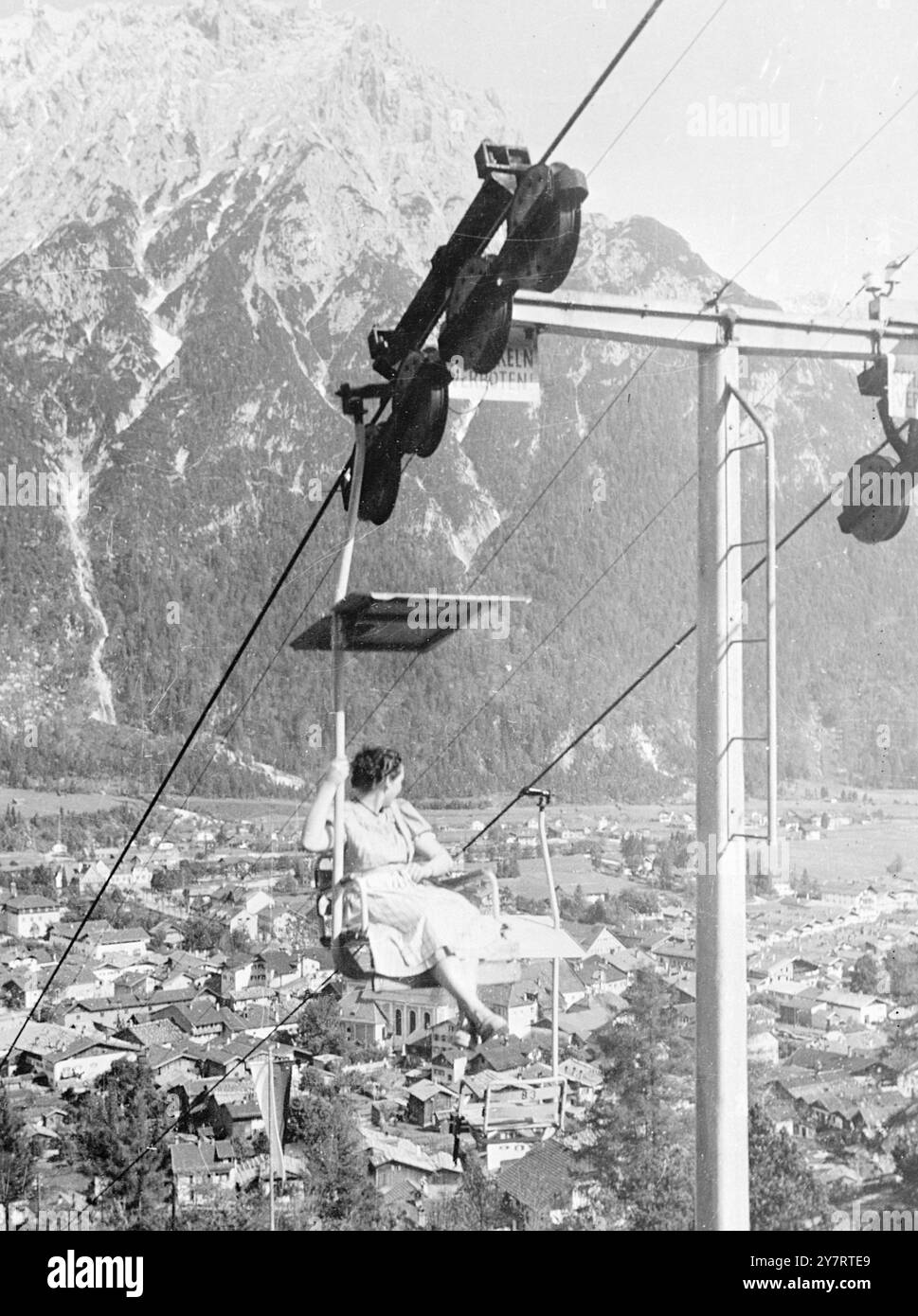 CAVALCA MITTENWALD 20.7,53. Tre anni fa, il villaggio tedesco di Mittenwald ha installato una seggiovia sulla cima di Kranzberg alta 1392 metri. Da allora, più di 300.000 passeggeri hanno goduto della vista dai 1450 metri di lunghezza. La foto mostra una splendida vista dalla seggiovia fino alla cima di 1392 metri di Kranzberg, ma la donna passeggero trova confortante tenersi al bar mentre guarda i tetti così in basso. 20 luglio 1953 Foto Stock