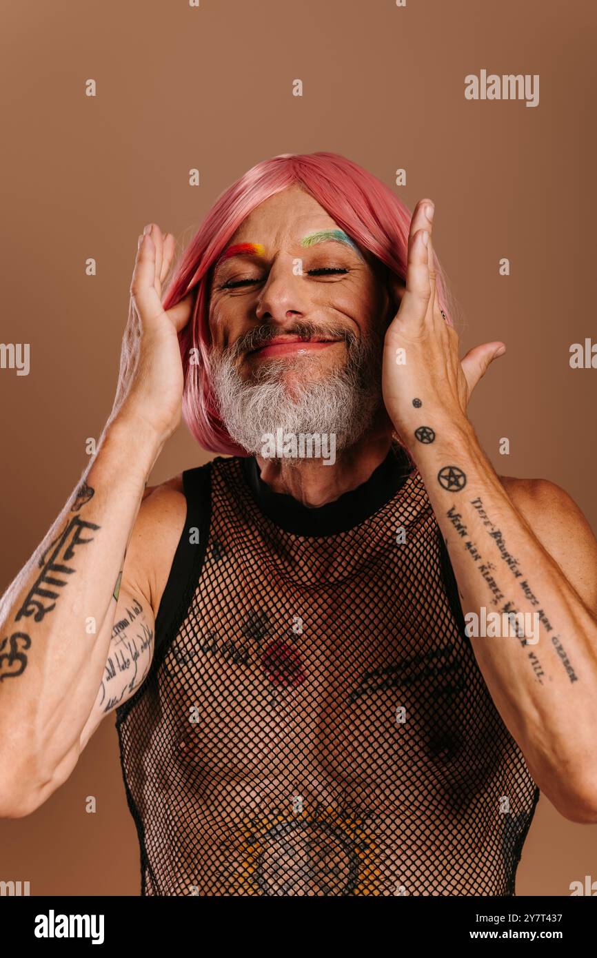 Felice uomo gay maturo con sopracciglia color arcobaleno che regola la parrucca rosa su sfondo marrone Foto Stock