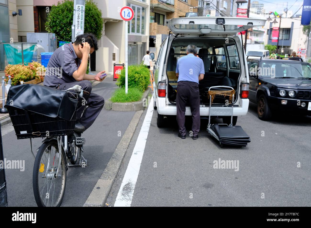 Un messaggero di biciclette seduto sulla sua bicicletta e guardando il suo telefono quando si prende una pausa con il personale addetto alla consegna e il suo furgone sullo sfondo. Harajuku, Shibuya Ward, Tokyo Foto Stock