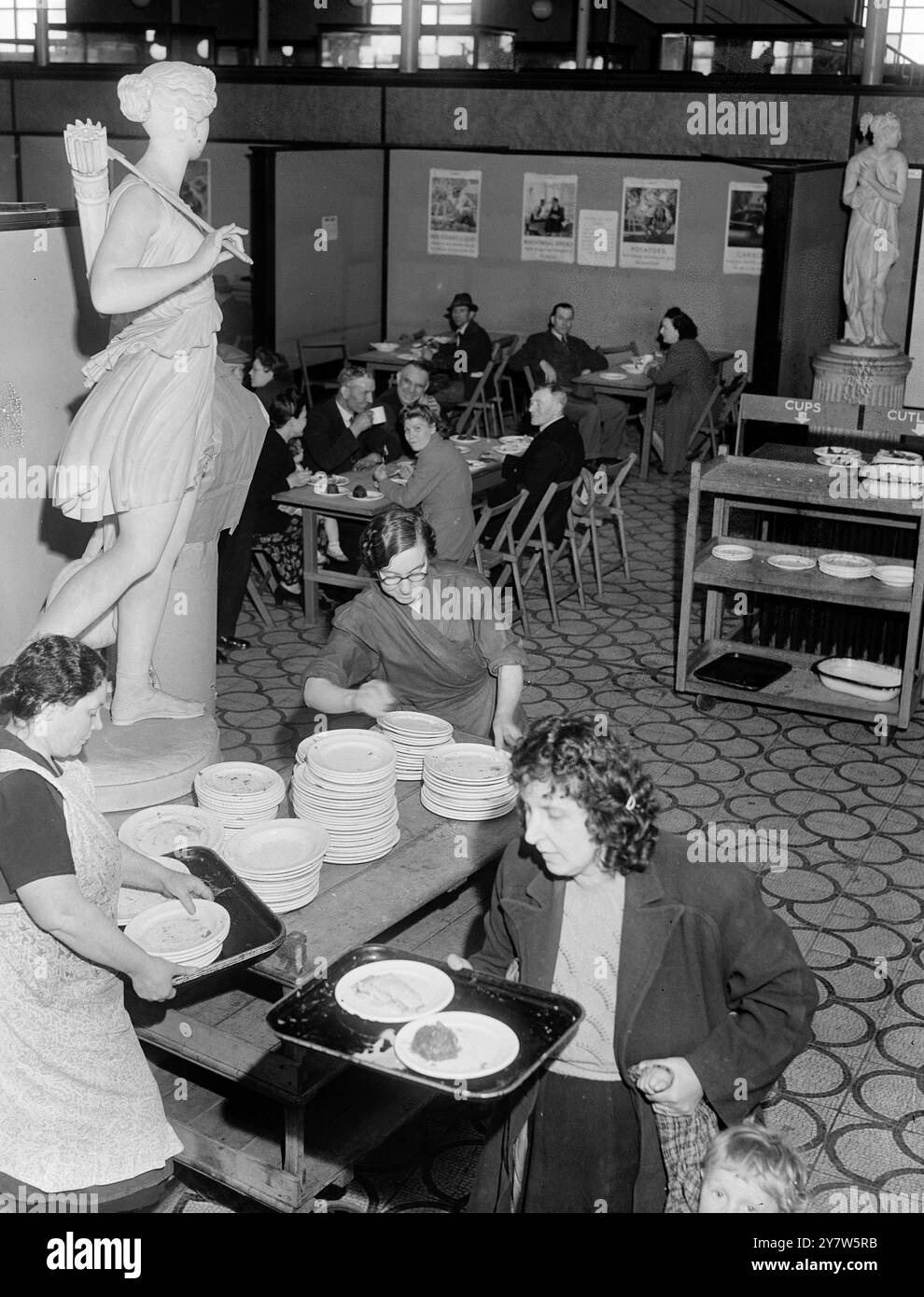 LONDINESI PRANZO CON GLI DEI: PASTI DA MUSEO A PREZZI POPOLARI ristoranti britannici, aperti dal Londoners' Meals Service per fornire buon cibo a prezzi minimi, sono apparsi quasi ovunque a Londra, da Park Lane a Putney, durante la guerra. Il più strano è al Bethnal Green Museum, nell'East End, dove ogni giorno possono essere serviti 1000 pranzi caldi. I clienti pagano dieci pence o uno scellino nel salone, in uno stand accanto a un busto di Garibaldi, il patriota italiano, e fanno il loro pasto presso un banco di servizio, portandolo davanti a un busto di Oliver Cromwell, Lord Protettore del commonwealth, al c Foto Stock