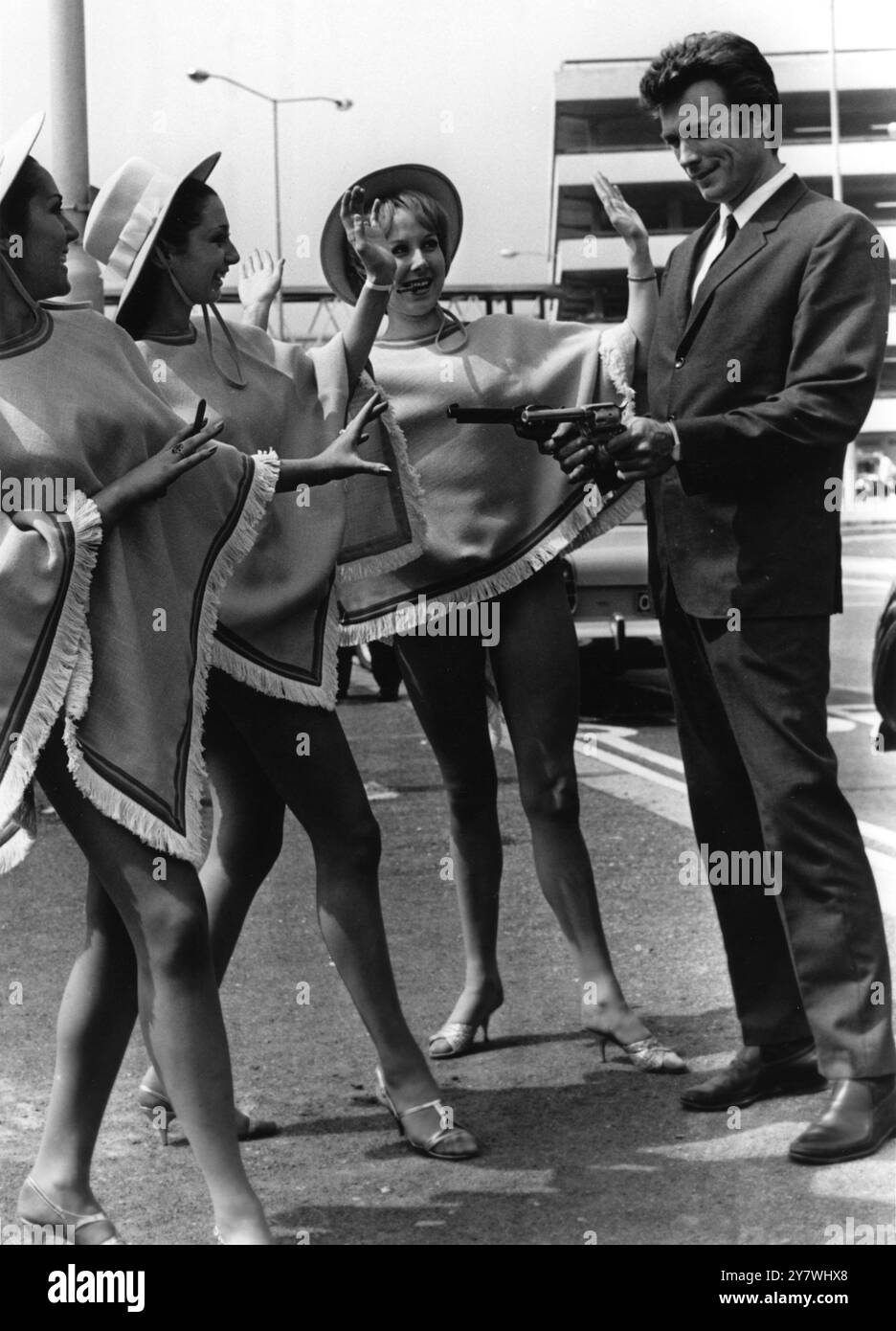 Clint Eastwood punta le sue pistole alle tre ragazze accoglienti vestite con mini outfit in stile squaw all'aeroporto di Heathrow a Londra. Clint Eastwood era in Gran Bretagna per promuovere il suo ultimo film, "Un pugno di dollari". 1 giugno 1967 Foto Stock