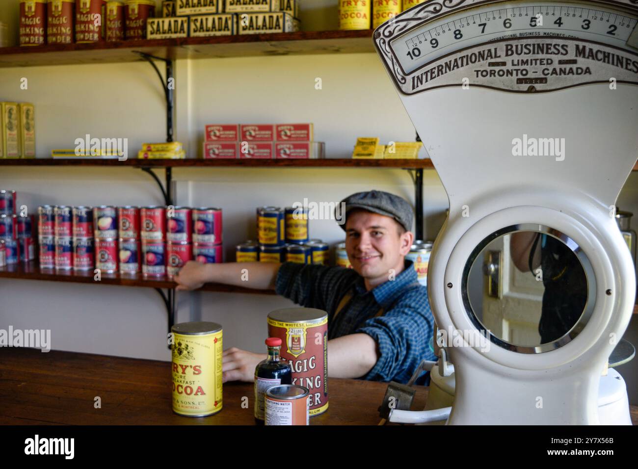 Un uomo vestito con abiti vintage si trova dietro il bancone di un vecchio negozio, mostrando abbigliamento storico e pratiche commerciali tradizionali. Foto Stock
