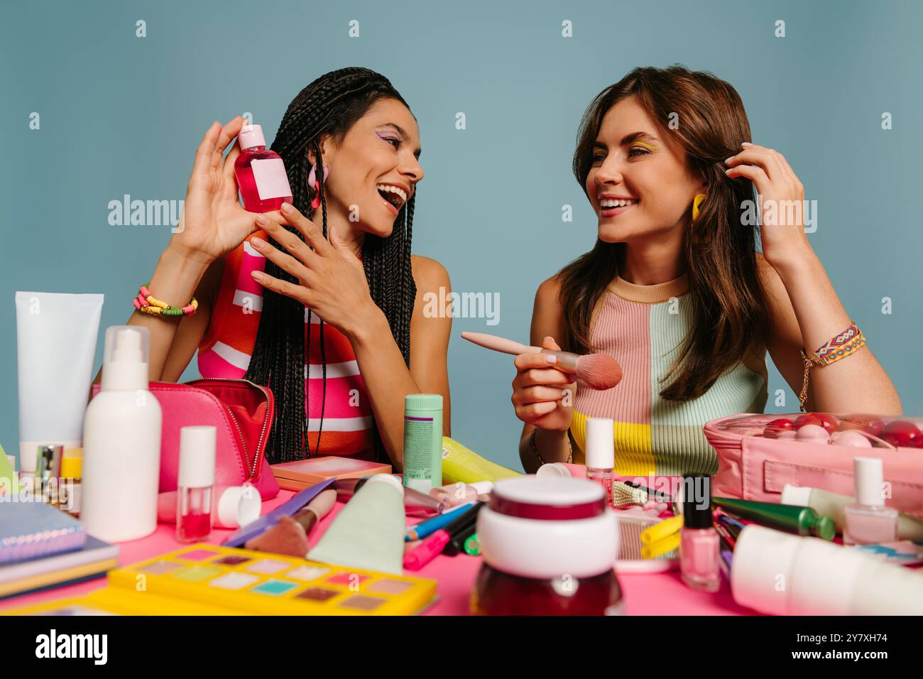 Due influencer donne felici che esaminano vari prodotti di bellezza mentre sono sedute alla scrivania su sfondo blu Foto Stock