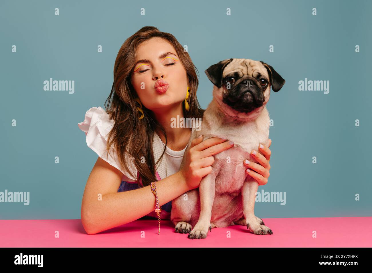 Bella donna che soffia come bacio e abbraccia un carino cane mentre siede alla scrivania rosa su sfondo blu Foto Stock